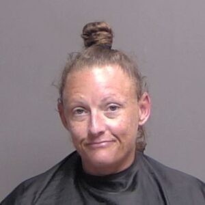 A woman with light skin and reddish brown hair in a topknot, wearing a black drape, stands against a plain gray background, looking at the camera A woman with light skin and reddish brown hair in a topknot, wearing a black drape, stands against a plain gray background, looking at the camera