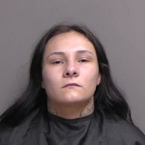 A woman with long dark hair, a neutral expression, and neck tattoos wears a black garment against a plain background
