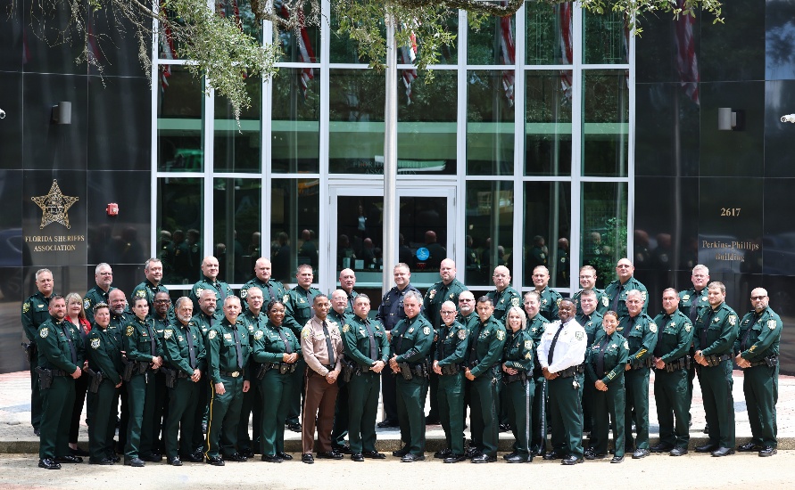 A group of uniformed law enforcement officers pose for a photo outside a building with glass doors and a sign reading “Florida Sheriffs Association ” A group of uniformed law enforcement officers pose for a photo outside a building with glass doors and a sign reading “Florida Sheriffs Association ”