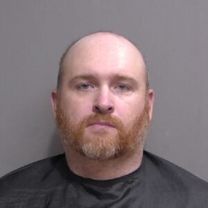 A man with a shaved head and red beard faces forward, wearing a black drape, against a plain gray background A man with a shaved head and red beard faces forward, wearing a black drape, against a plain gray background