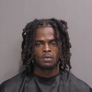 A mugshot of a young Black man with locs, a light beard, and a serious expression, wearing a black shirt against a plain grey background A mugshot of a young Black man with locs, a light beard, and a serious expression, wearing a black shirt against a plain grey background