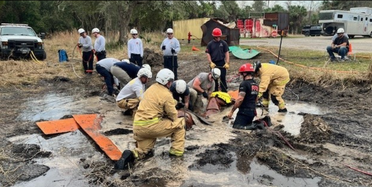 Rescue workers in helmets and gear work together in a muddy area to assist a horse that is trapped, using straps and boards for support, while others observe and offer help nearby