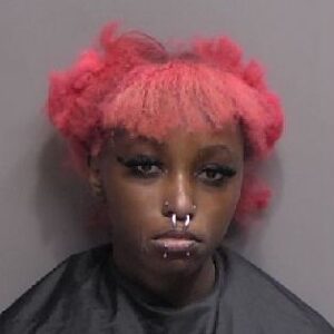 A person with bright pink hair, multiple facial piercings, and dramatic eye makeup is wearing a dark cape and looking straight at the camera against a plain background
