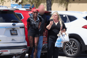 A person with red hair is being escorted by a man while another woman walks beside them in a parking lot The person appears upset, and the group is surrounded by parked cars