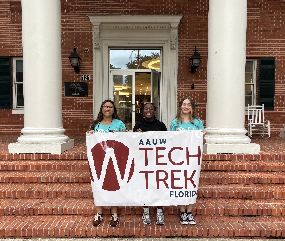 Three young women stand on brick steps in front of a building holding a banner that reads, “AAUW Tech Trek Florida ”