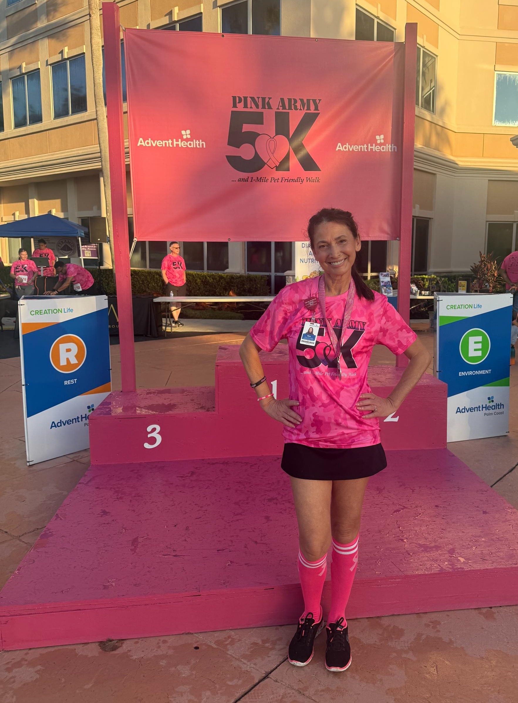 A woman wearing a pink race t shirt and finisher medal stands on a pink race podium in front of a Pink Army K sign