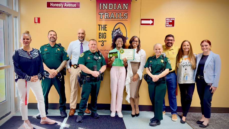 A group of people, including students, teachers, and police officers, pose indoors One student holds a certificate Behind them are yellow walls, signs, and a banner reading “Indian Trails” and “The Big ”