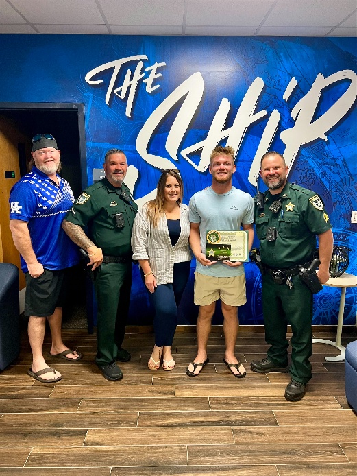 Five people pose indoors; three wear sheriff uniforms, and two are in casual clothes One person holds a certificate and a plaque A blue wall behind them features large text reading “The Ship ”