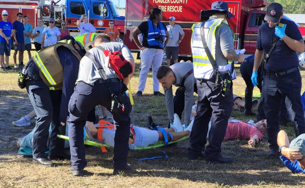 Emergency responders attend to people lying on the ground during a drill or rescue scenario, with fire trucks and onlookers in the background Some responders are kneeling and assisting a person on a stretcher